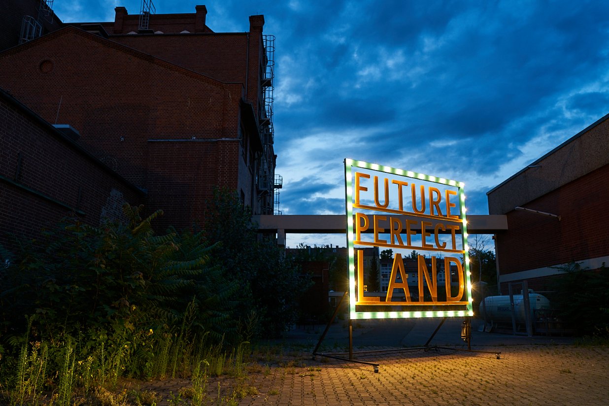 Ein Schild mit der Schrift &quot;FuturePerfectLand&quot; steht auf einem Industriegel&auml;nde, das teilweise von Pflanzen &uuml;berwachsen ist, unter einem drohenden Himmel. | © Pablo Hassmann, Magma Studio