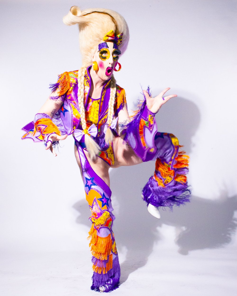 Cherdonna Shinatra in buntem Drag Kost&uuml;m, mit clowneskem Drag MakeUp, hebt das linke Bein, den linken Arm und guckt erstaunt. | © Stephen Anunson